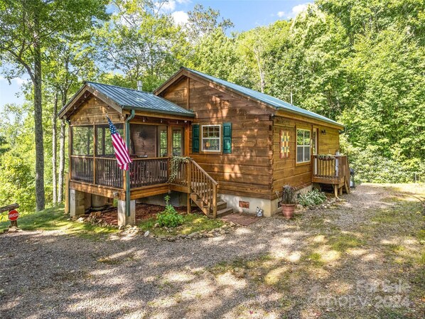 Une cabane en bois avec un porche, un drapeau et une allée de gravier, le tout entouré d’arbres.