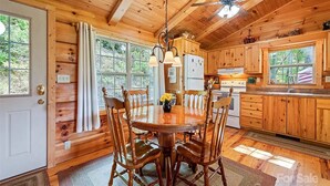 Interior de una cabaña de madera con una mesa de comedor rodeada de sillas, una zona de cocina con armarios de madera y un ventilador de techo.