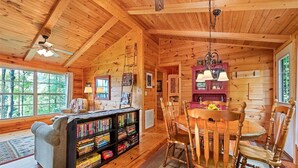 Acogedor interior de cabaña con techo de madera, chimenea, comedor con sillas de madera y una estantería llena de libros.