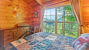 Un dormitorio de cabaña de madera con un ventanal grande, una cama con colcha, un aparador y un espejo en la pared.