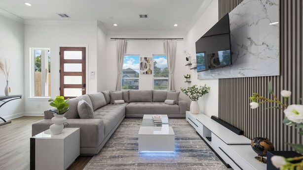 A modern living room with a sectional sofa, a coffee table, a flat-screen TV mounted on a marble wall, and a large window with curtains.