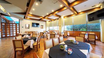 A restaurant with multiple tables and chairs, a TV mounted on the wall, and large windows.