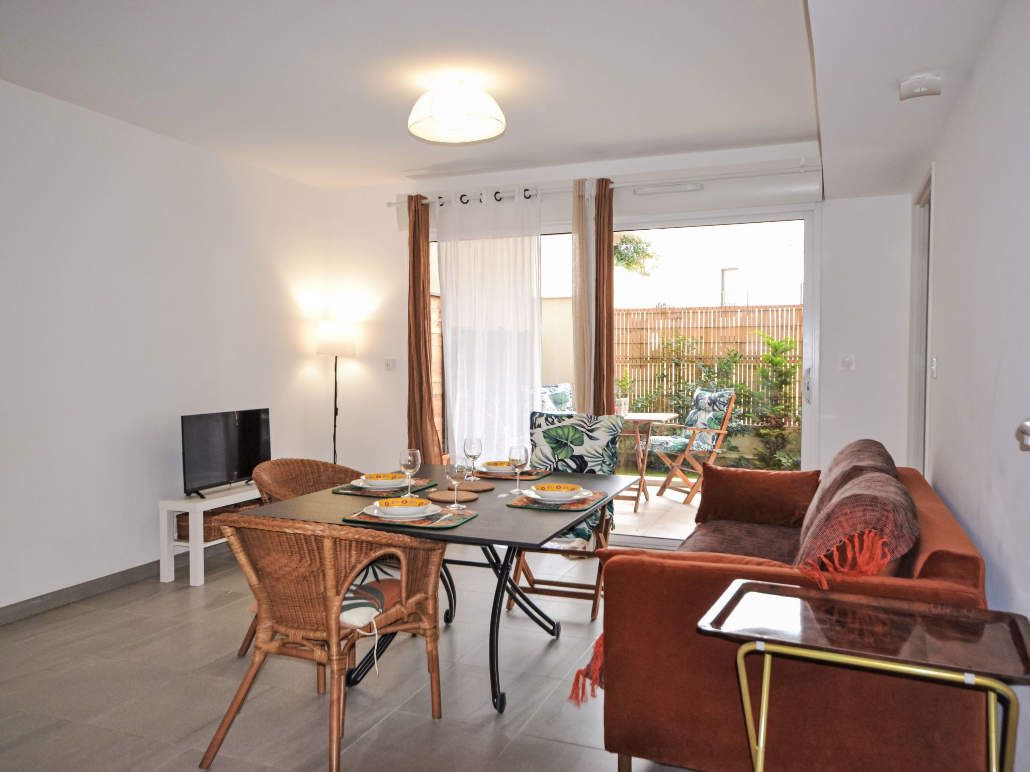 Un coin repas avec une table dressée pour quatre personnes, un canapé, des fauteuils en osier et un téléviseur posé sur un meuble.