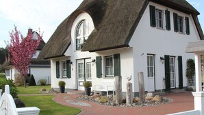 A white house with a thatched roof, green shutters, and a brick pathway leading to the entrance.