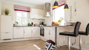 A modern kitchen with white cabinets, stainless steel appliances, a dining area with two chairs, and a window with striped curtains.