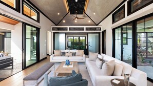Ein modernes Wohnzimmer mit einem weißen Sofa, einer Bank und einem Couchtisch. Ein Deckenventilator und große Fenster sorgen für natürliches Licht.