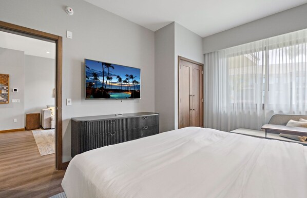 A modern hotel room with a large bed, a flat-screen TV mounted on the wall, a wooden wardrobe, and a large window with sheer curtains.