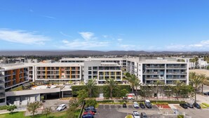 A modern multi-story building with a parking lot in front, surrounded by palm trees and a clear sky.