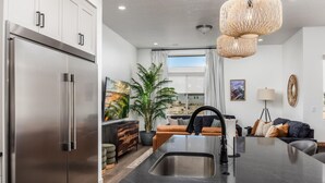 A modern kitchen with a stainless steel refrigerator, a black countertop, and a sink. There is a dining area with a sofa and a plant. - Adventure Central at Desert Color (by Ember Stays) (St. George)