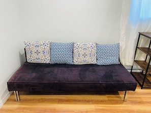 A dark-colored sofa with four patterned cushions against a plain wall, a wooden floor, and a metal shelf unit to the right.
