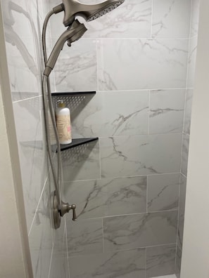 A shower with a handheld showerhead, a bottle of shampoo, and marble-patterned tiles.
