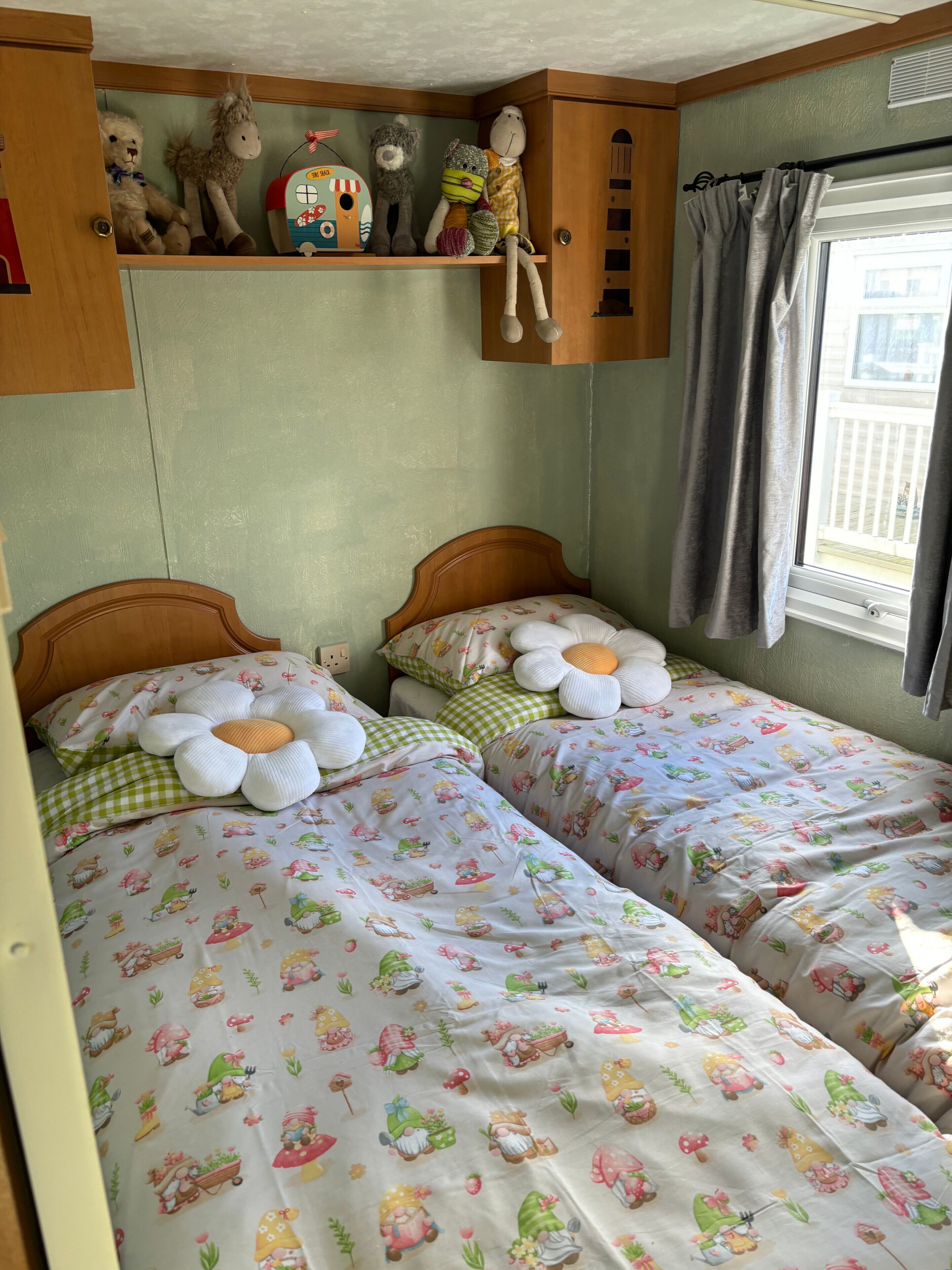 A cozy bedroom with two beds, each with floral bedding and decorative pillows. A shelf above the beds holds various plush toys and a toy train.