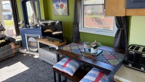 Un espacio habitable compacto con una televisión, una mesita con camino de mesa decorativo, una ventana con cortinas y una repisa con una figurita de pájaro.