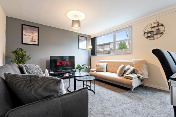 Una sala de estar moderna con un sofá gris, un sillón negro, una mesa de centro de vidrio y un televisor de pantalla plana que muestra 'NETFLIX'.