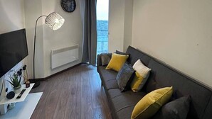 A modern living room with a sofa, a TV, a floor lamp, a wall clock, and a window with curtains. - 1BR-Sleeps4-Parking-ClosetoCentre (Birmingham)