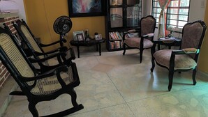 A living room with two wooden chairs, a small table with a fan, a bookshelf, and a painting on the wall.