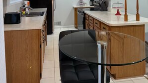 A modern kitchen with wooden cabinets, a black refrigerator, a glass dining table with black chairs, and a unique pendant light. - Charming 3-bedroom house in peaceful White Lake Township on Pontiac Lake (White Lake Township)