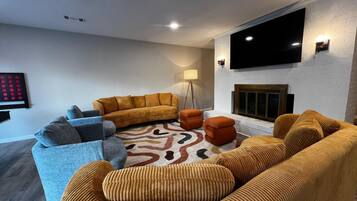 Un salon moderne avec un grand canapé, un canapé plus petit et deux poufs. Un téléviseur à écran plat est fixé au mur, et une cheminée se trouve en dessous.