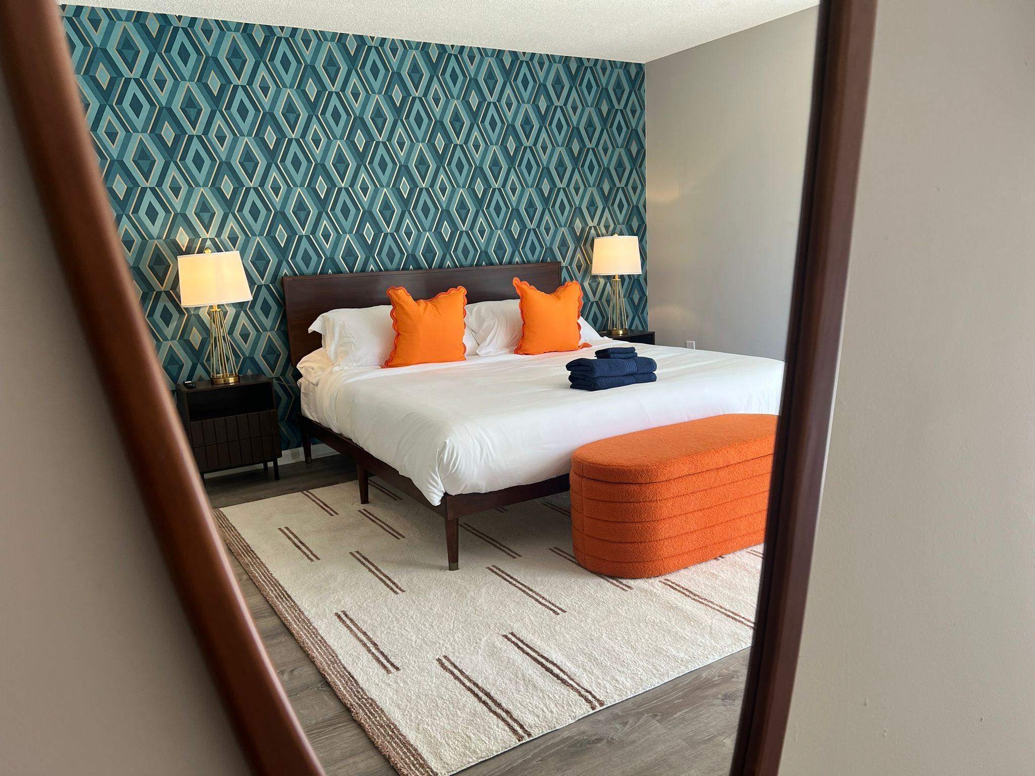 Un lit bien fait, avec une literie blanche et orange, un pouf assorti de la même couleur et un mur à motifs en arrière-plan.