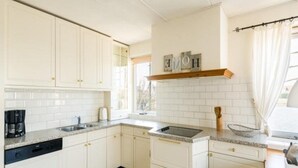 Una cocina con armarios blancos, un lavavajillas negro, una cafetera y un ventanal con cortinas.
