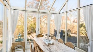 Un invernadero con una mesa y sillas de madera, una mesita auxiliar y vistas al exterior.