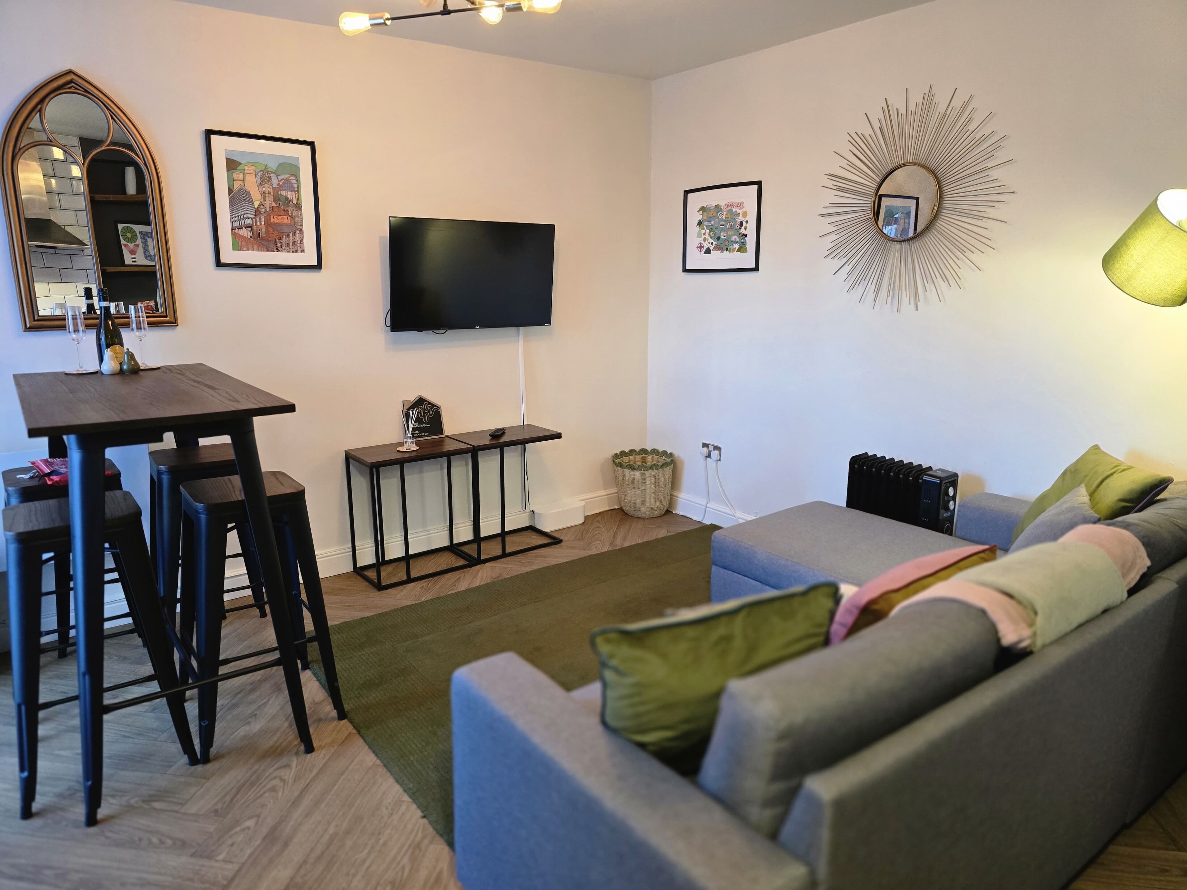 Een moderne woonkamer met een grijze bank, een kleine eettafel met krukken, een flatscreen televisie en decoratieve wandkunst.