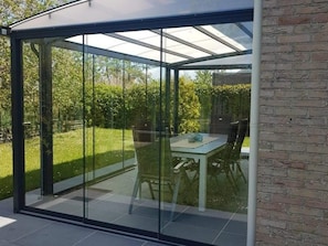 Une terrasse fermée par des parois de verre, avec une table et des chaises, entourée d’un mur de briques et d’une pelouse.