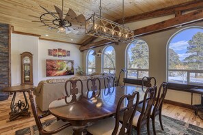 Un comedor con mesa y sillas de madera, una araña y ventanas amplias con vistas a árboles.