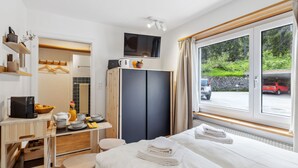 A hotel room with a bed, a desk with a computer, a television, and a window with a view of a parking area and greenery. - Promenade Apt 016 by Interhome (Arosa)