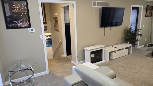 A living room with a white sofa, a glass coffee table, a fireplace, a TV mounted on the wall, and a door leading to another room.