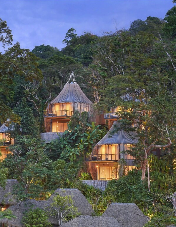 A cluster of treehouse-style accommodations nestled among dense foliage, featuring multiple levels and a thatched roof.