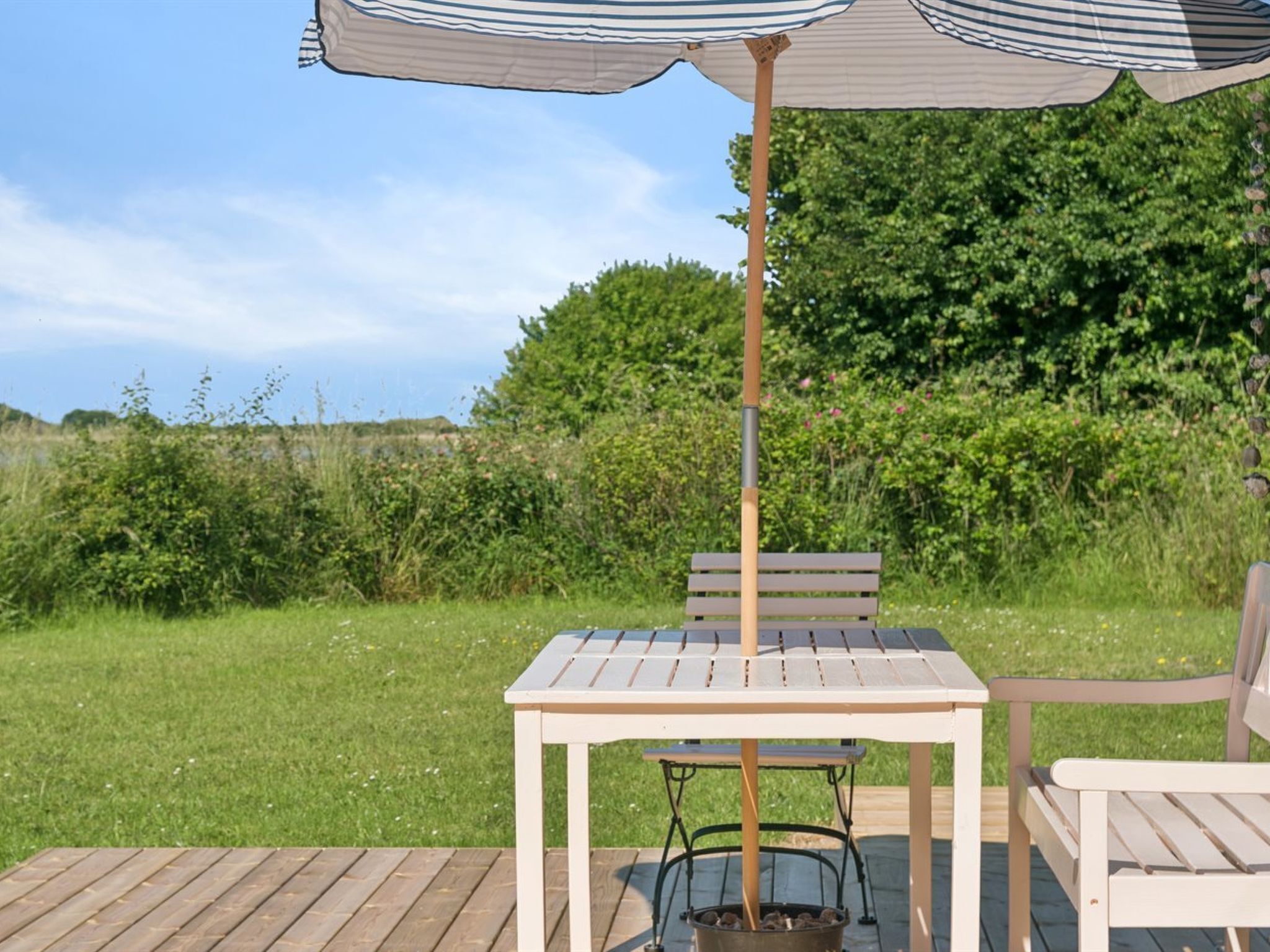Eine Holzterrasse im Freien mit Tisch, Stühlen und einem gestreiften Sonnenschirm, sowie einem Metalltopf.