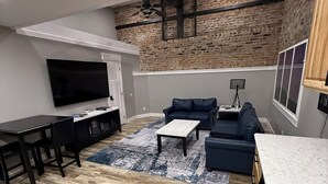 Ein modernes Wohnzimmer mit Flachbildfernseher, dunklen Sofas, einem Couchtisch und einer Küchenzeile mit Backsteinwand.