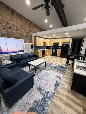 A modern living room with a black leather sofa, a coffee table, and a large rug. There is a kitchen in the background with wooden cabinets and a black refrigerator.
