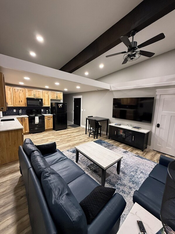A modern living room with a black leather sofa, a coffee table, and a TV. The kitchen is visible in the background with wooden cabinets and a black refrigerator.