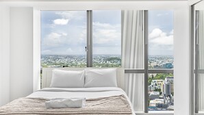 Een hotelkamer met een groot bed, witte beddengoed en uitzicht op een stadsgezicht door grote ramen.