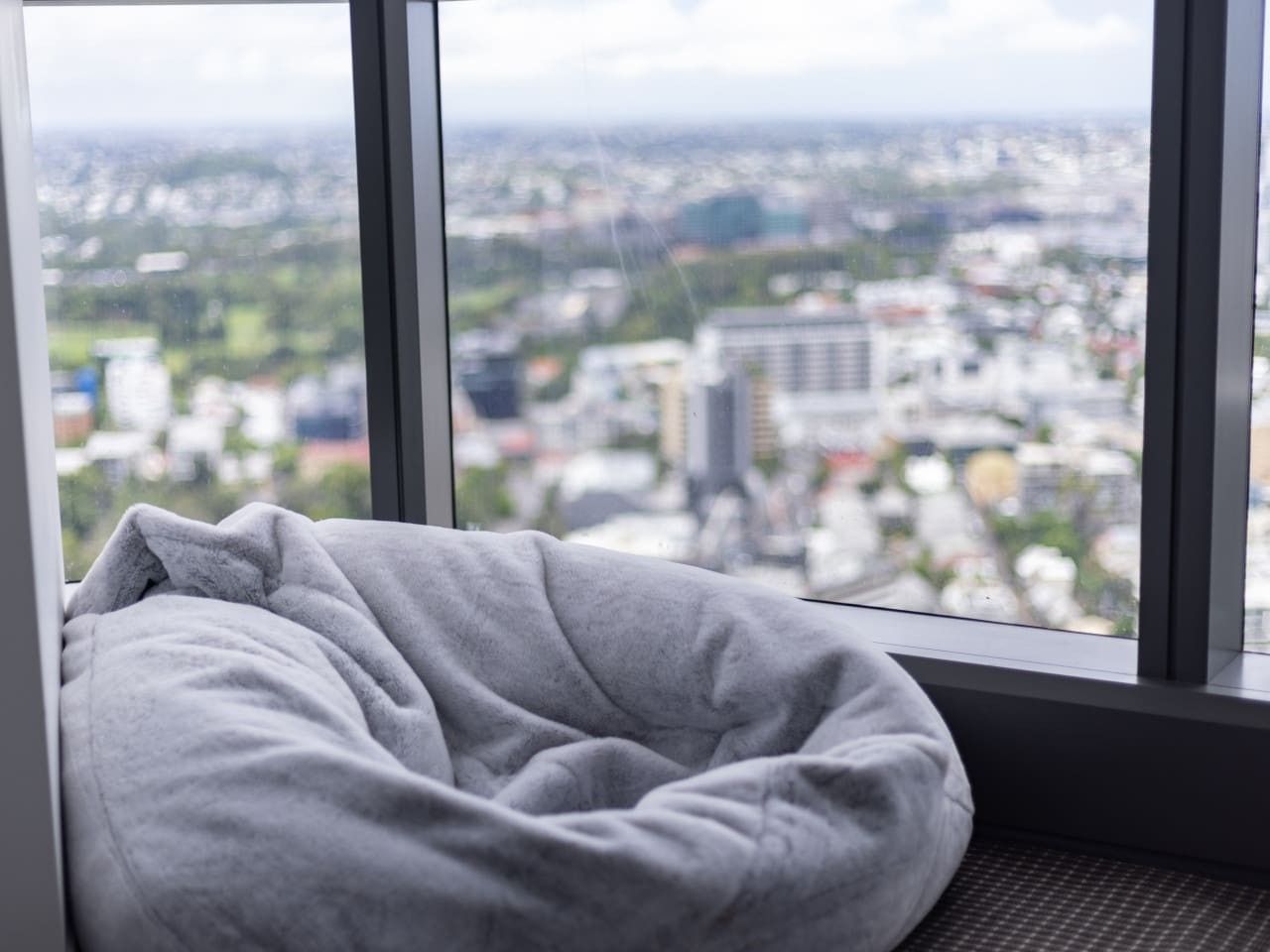 A window seat with a gray cushion, overlooking a cityscape with buildings and greenery.