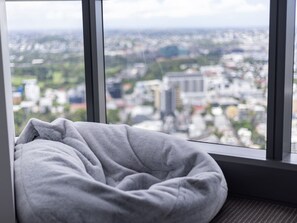 Asiento junto a la ventana con un cojín gris, con vistas a un paisaje urbano con edificios y zonas verdes.