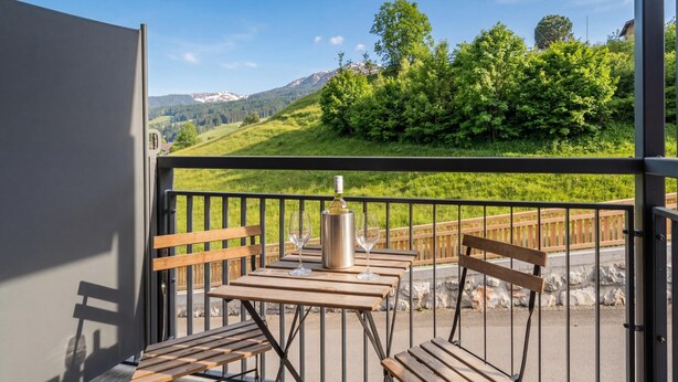 Ein Balkon mit Tisch und Stühlen, einer Weinflasche und Blick auf eine grüne Landschaft mit Bäumen und Bergen.