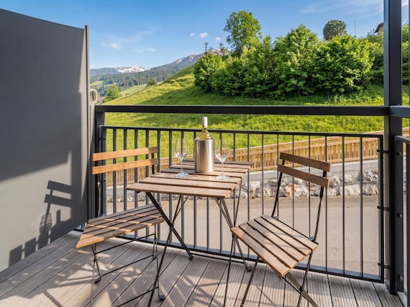 A balcony with a table and chairs, a wine bottle, and a view of a green landscape with trees and mountains.