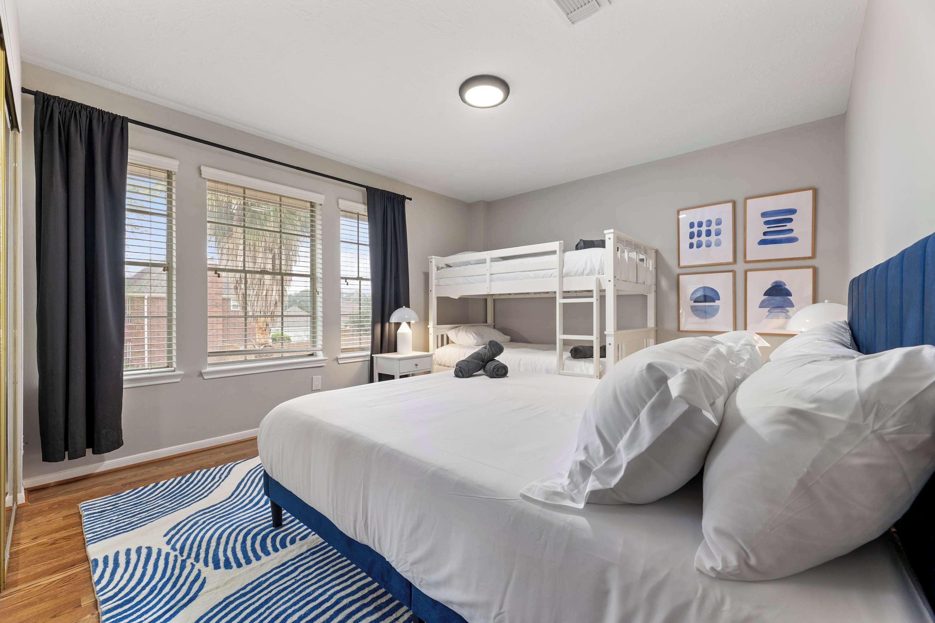 Een slaapkamer met een stapelbed, een groot bed, een blauw-wit gestreepte vloerkleed en een raam met zonwering.