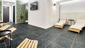 Een moderne spa ruimte met een vloer met zwarte stipjes, witte bakstenen muren en een flatscreen televisie aan de muur. Er staan houten ligbedden en een klein tafeltje met stoelen.