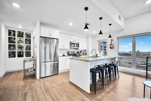 Una cocina moderna con armarios blancos, electrodomésticos de acero inoxidable, una isla grande con taburetes y vistas a la ciudad a través de amplios ventanales.