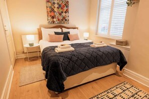 A neatly arranged bedroom with a bed, bedside lamps, a window with blinds, a decorative wall piece, and a small side table.