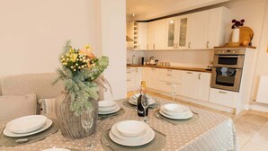 A dining area with a table set for four, a vase with flowers, and a kitchen in the background. - Chester coach house with log burner & parking (Cheshire West and Chester)