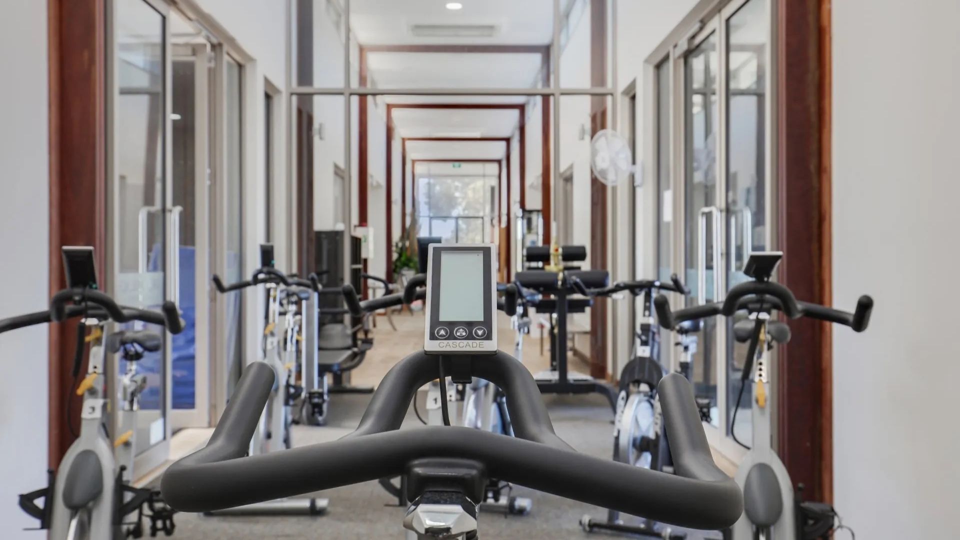 A gym with stationary bikes and cardio equipment, a digital display, and a corridor with mirrors and windows.