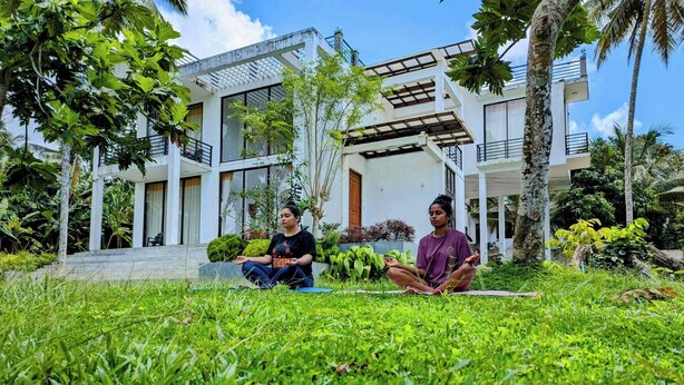 Two people meditating on a grassy lawn in front of a modern house with a balcony and large windows. - Room in Guest Room - Serene Oasis in Galle - Tranquil Retreat With Breathtaking Views (Galle)