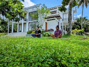 Two people meditating on a grassy lawn in front of a modern house with a balcony and large windows. - Room in Guest Room - Serene Oasis in Galle - Tranquil Retreat With Breathtaking Views (Galle)