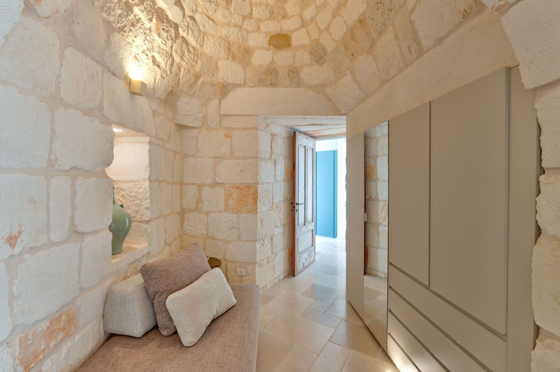 Un pasillo de paredes de piedra con un sofá y cojines, que conduce a una habitación luminosa con una pared azul.