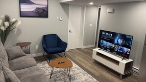 A modern living room with a grey sofa, a blue armchair, a wooden coffee table, and a flat-screen TV on a white stand. - 2-Bedrooms | On site Parking | near YQR Airport (Regina)